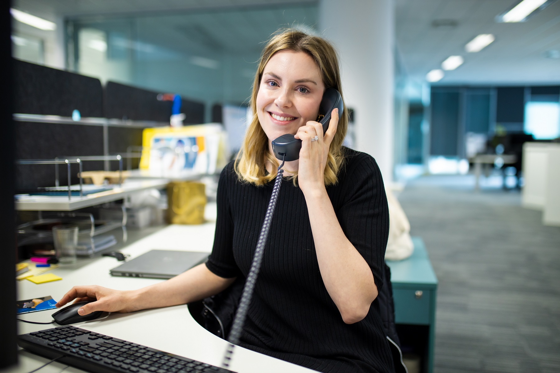 Image of a woman smiling at the camera, on the phone sitting behind a workstation and computer.